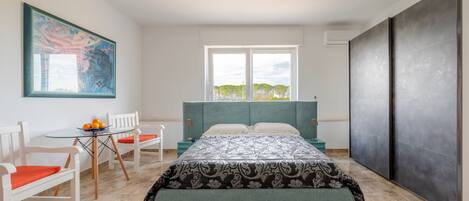 A bedroom with a bed, a window, a painting, a table with chairs, and a view of trees.
