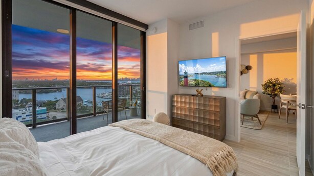Ein Schlafzimmer mit einem großen Bett, einem an der Wand montierten Fernseher, einer Schiebeglastür zu einem Balkon und Blick auf eine Stadtlandschaft bei Sonnenuntergang.