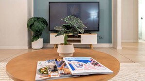 Een woonkamer met een ronde houten salontafel, een potplant en een tijdschrift. Een flatscreen televisie hangt aan de muur op de achtergrond.