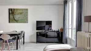 A modern living room with a flat-screen TV, a dining area with a table and chairs, and a large abstract painting on the wall. - Modern 3BR Apartment with Balcony - Pereire (Paris)