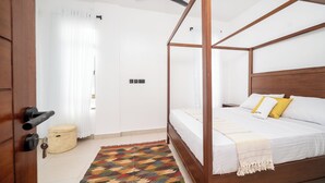 Un lit à baldaquin avec une literie blanche et un coussin jaune, un ventilateur de plafond, un panier en osier et un tapis coloré.