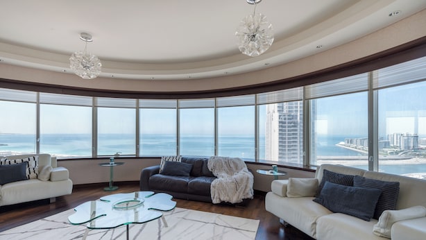 A spacious living room with a curved ceiling, large windows offering a view of the ocean and cityscape, a white sofa, a dark sofa, and a glass coffee table. - EDEN'S Homes & Villas - KG tower (Dubai)