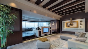 A modern living room with a large television, comfortable seating, a decorative wall unit, and a large window offering a city view. - EDEN'S Homes & Villas - KG tower (Dubai)