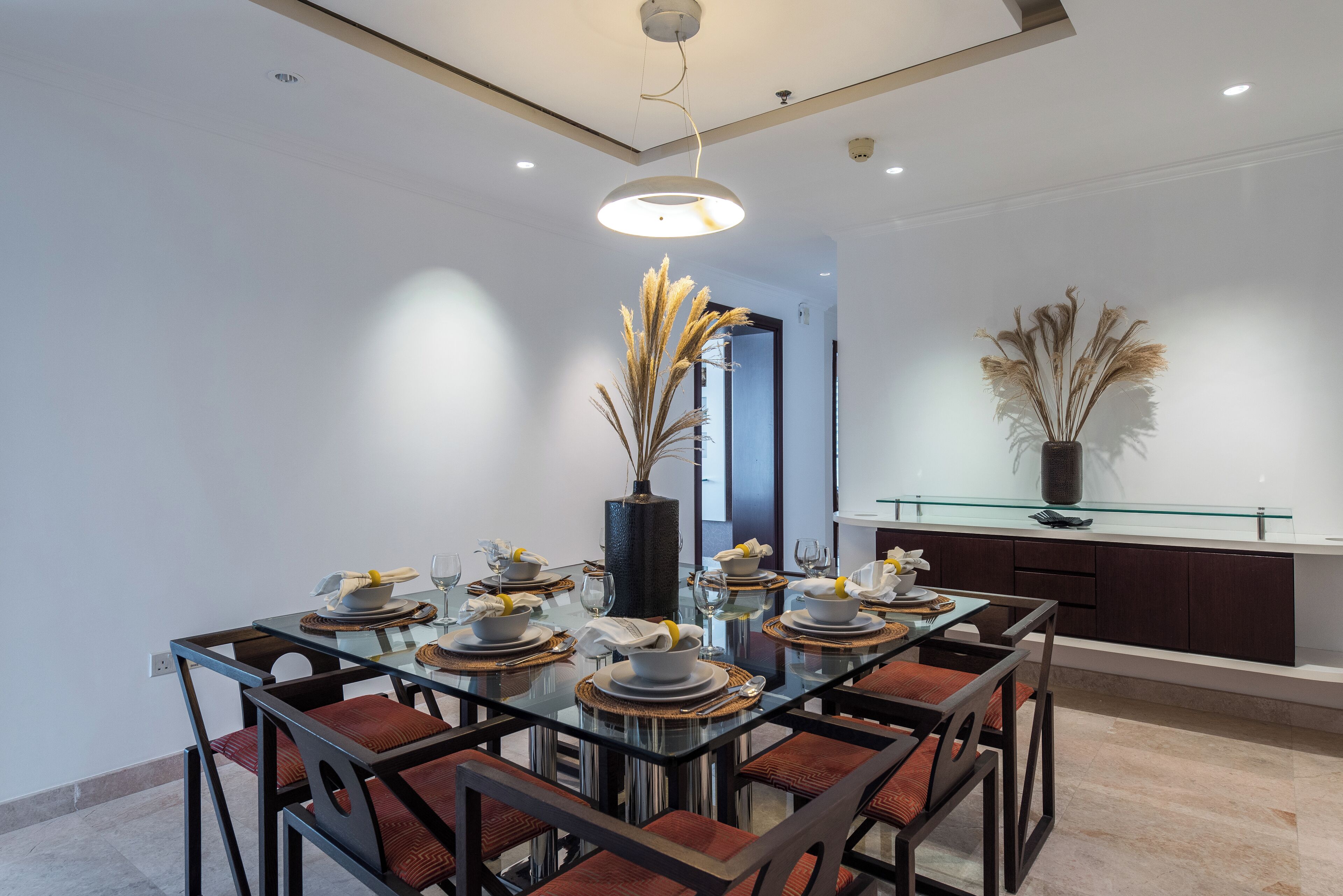Un comedor moderno con una mesa de vidrio, sillas oscuras y un jarrón con plantas secas.