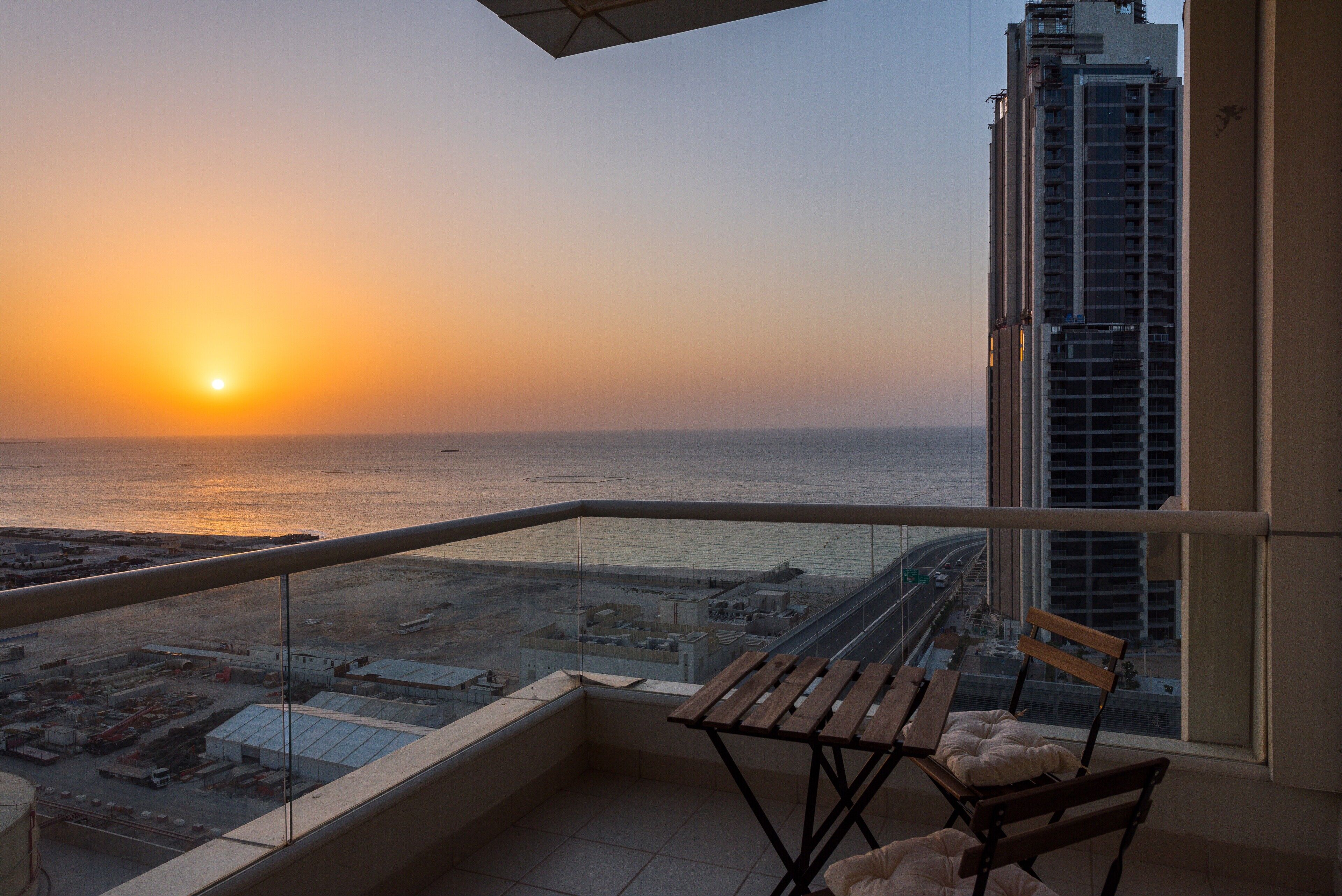 Un balcón con vista al atardecer sobre el mar, un edificio alto a la derecha y un pequeño rincón de descanso con cojines.