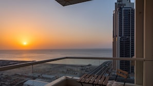 A balcony with a view of the sunset over the sea, a high-rise building to the right, and a small seating area with cushions. - EDEN'S Homes & Villas - KG tower (Dubai)