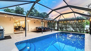 Una piscina con un área de terraza techada, una parrilla y vistas de los árboles circundantes.