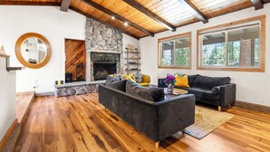 A spacious living room with a stone fireplace, wooden ceiling beams, and large windows offering views of the outdoors. - Steps to Trails & Minutes to Heavenly + Firepit | Bluewood Cabin by AvantStay (South Lake Tahoe)