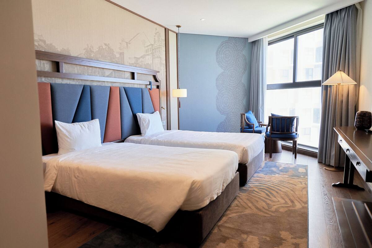 A hotel room with two beds, a desk, a chair, and a large window.