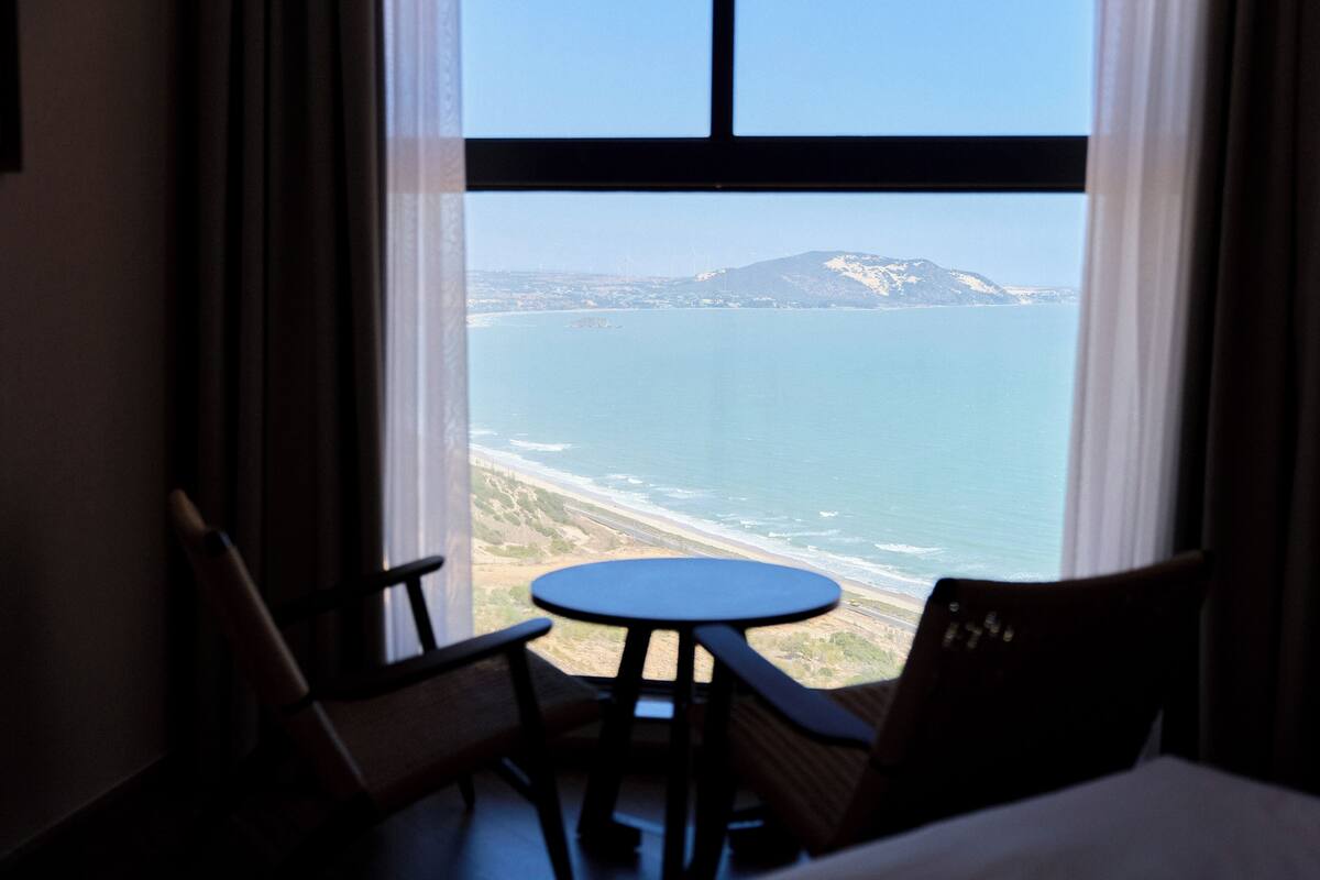 A room with a view of the ocean and a mountain. There is a table and two chairs.
