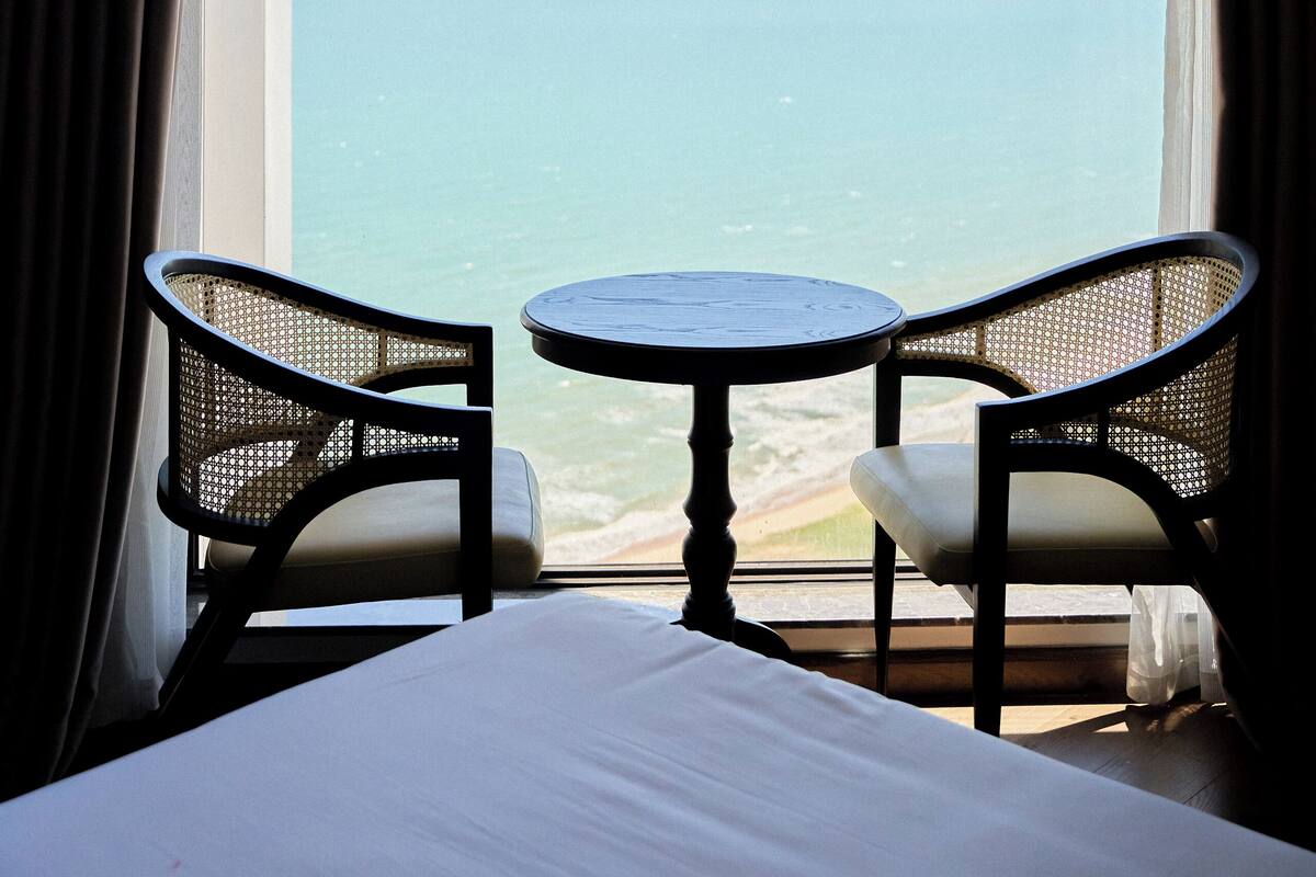 A hotel room with a large window, two wicker chairs, a small round table, and a bed.