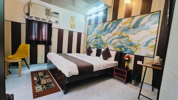A bedroom with a bed, a chair, a small table, a wall-mounted air conditioner, and a decorative painting. - Roomshala Hotel Mannat (New Delhi)
