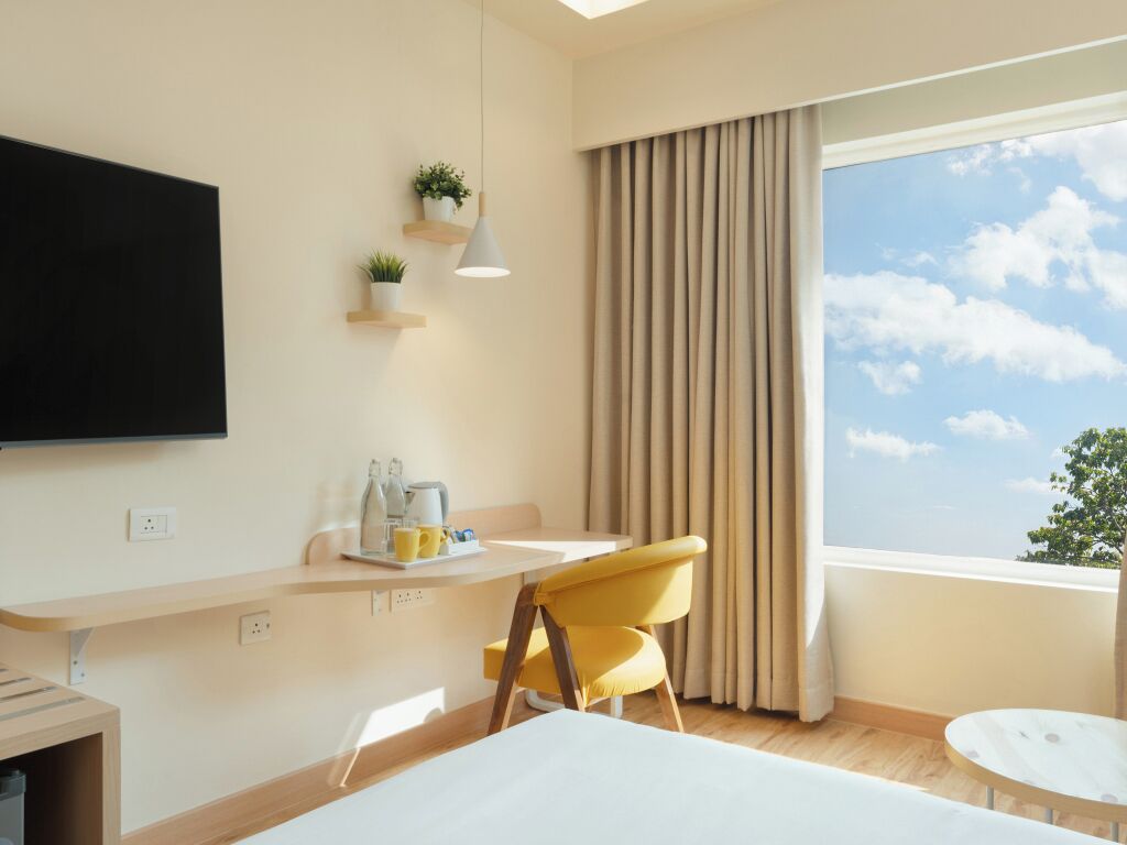 A modern hotel room with a large TV, a desk with a chair, a small table, and a view of the sky through the window.