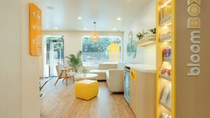 A modern living room with a yellow ottoman, a sofa, and a dining area with a yellow cabinet labeled 'bloomBox'. - Bloom Hotel - M Block GK2 (New Delhi)