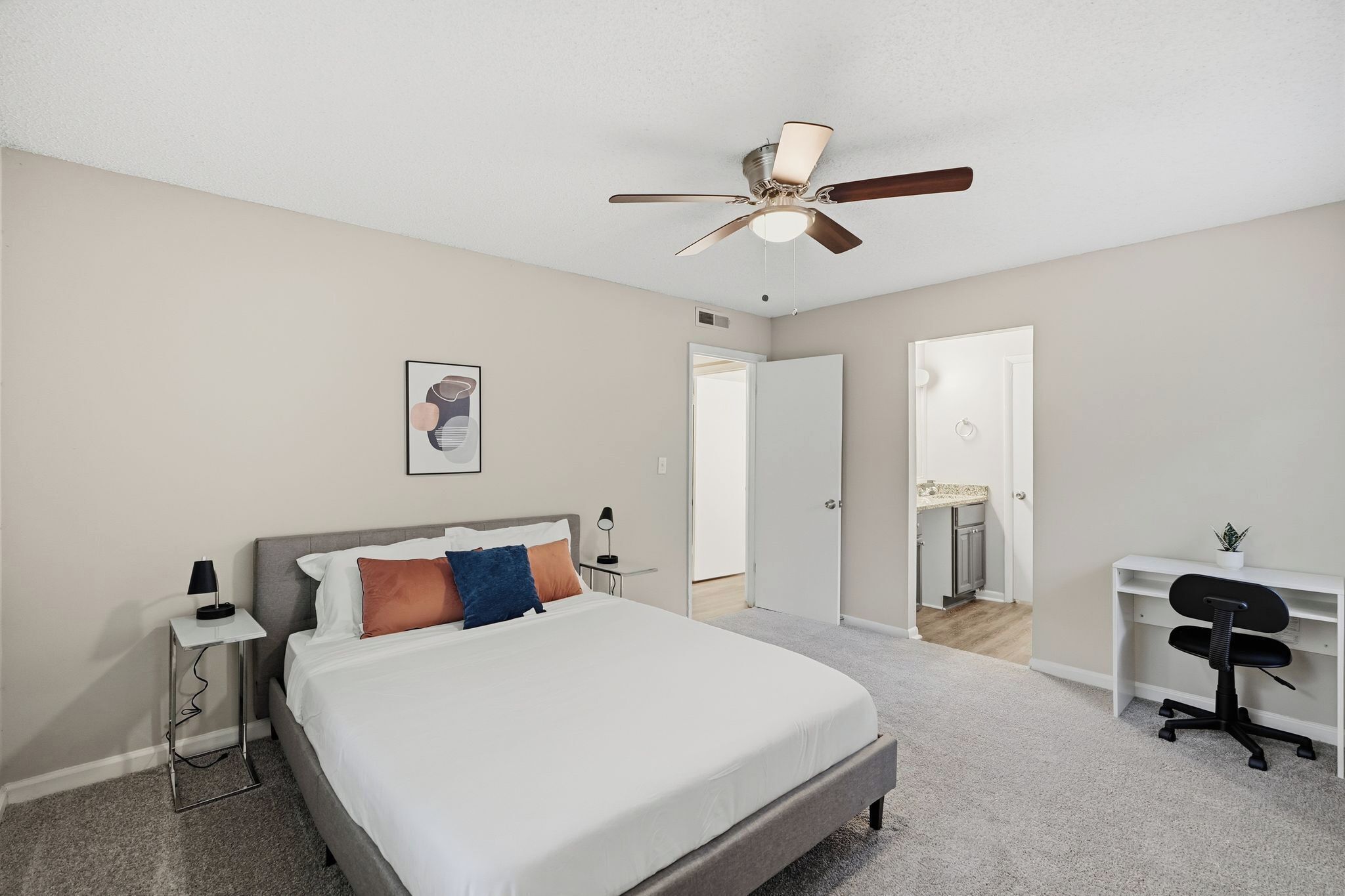 Une chambre à coucher comprenant un lit, un bureau, une chaise, un ventilateur de plafond et une porte donnant sur une salle de bain.