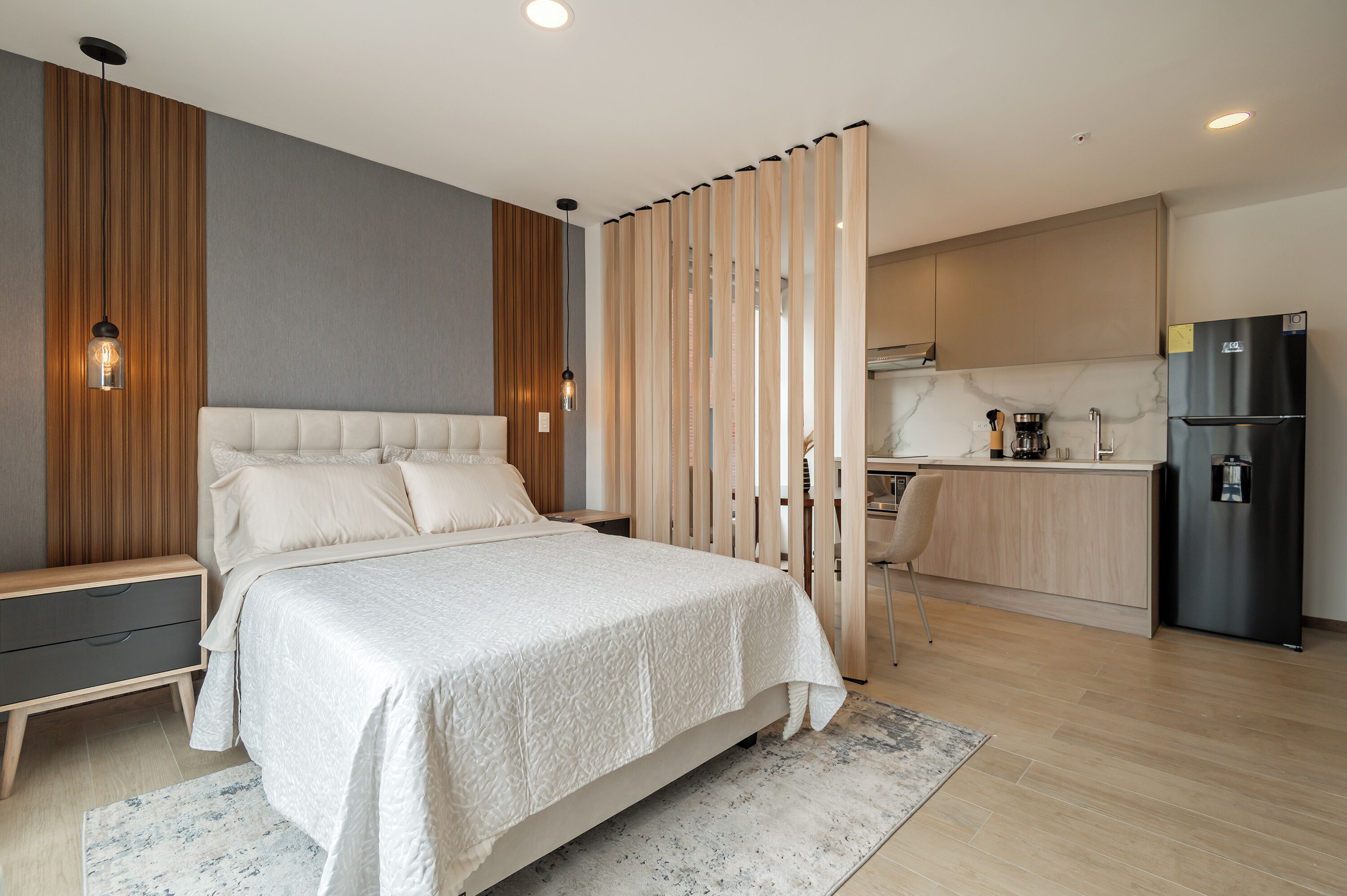 A modern bedroom with a large bed, a bedside table, and a nightstand. There is a kitchen area with a refrigerator and a coffee maker.