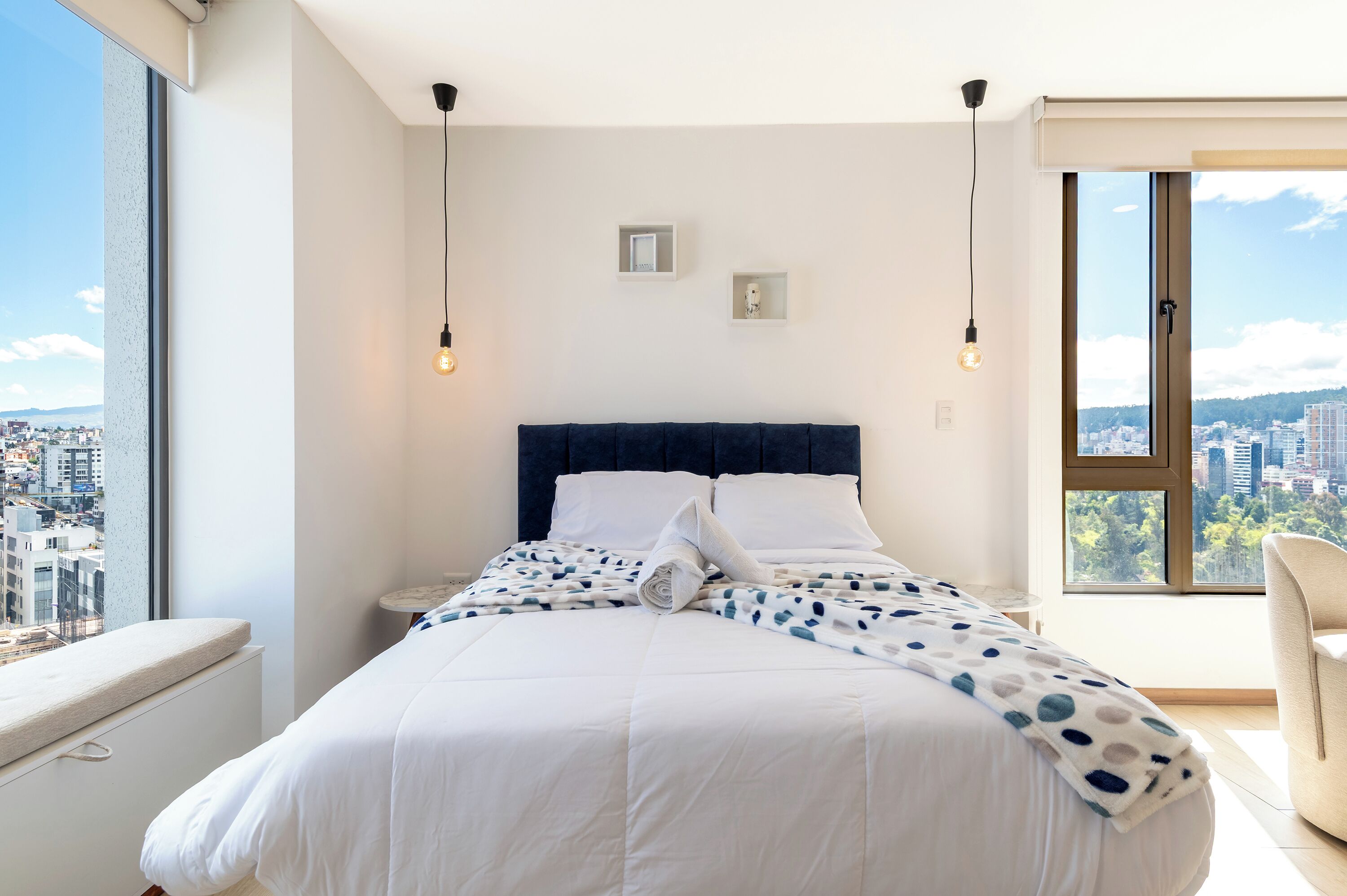 Una habitación de hotel moderna con una cama grande, vistas a la ciudad y una mesita de noche con lámpara.