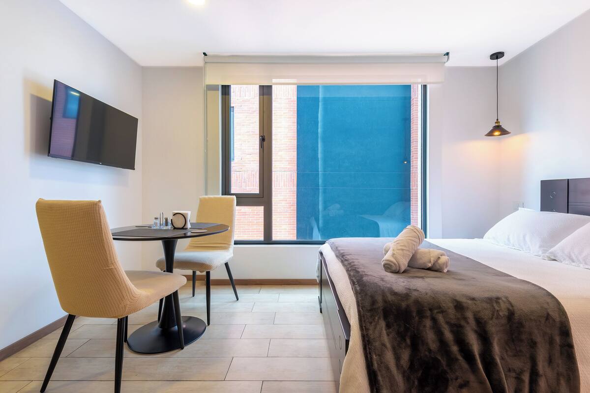 Una camera d'albergo moderna con un letto, una scrivania con sedia, una TV a parete e un'ampia finestra vista città.