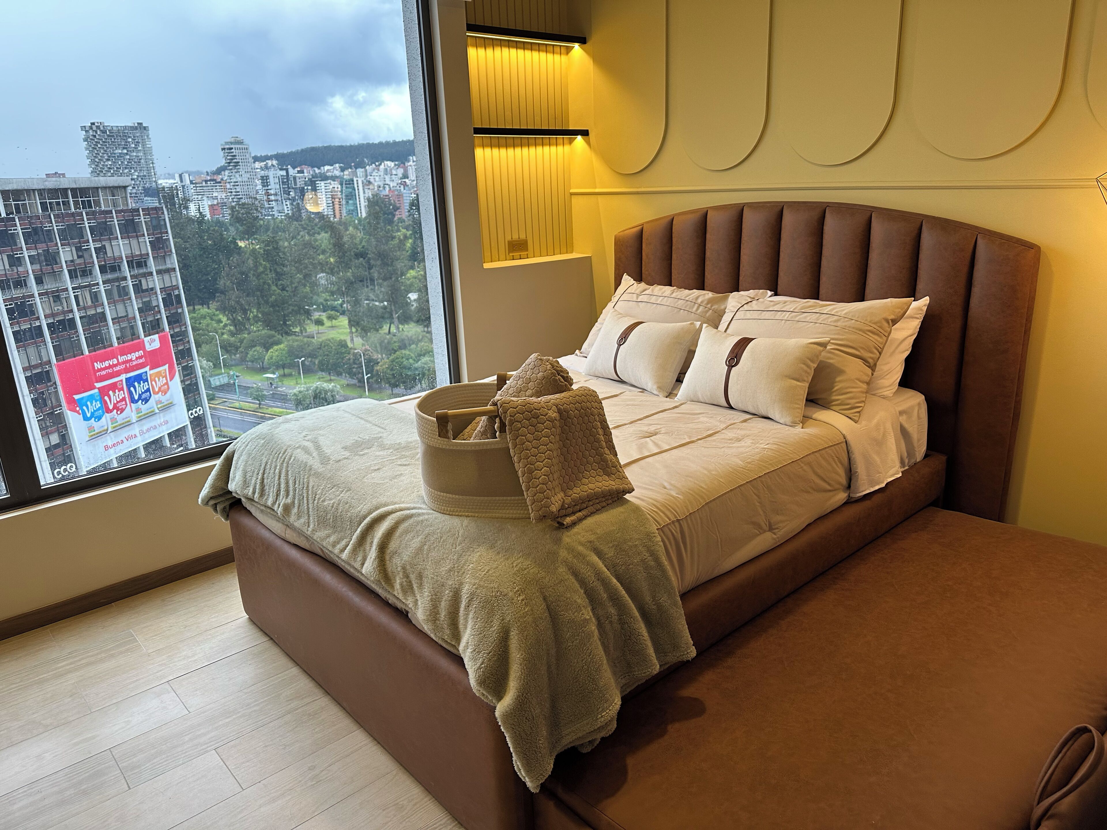 Ein modernes Schlafzimmer mit großem Fenster, einem Bett mit beigefarbener Bettwäsche und Blick auf eine Stadtlandschaft.
