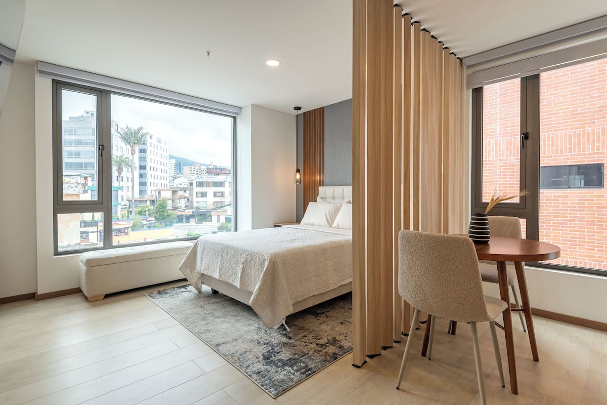 Una moderna camera d'albergo con un letto grande, un tavolino con una sedia, una finestra con tende e vista sulla città.