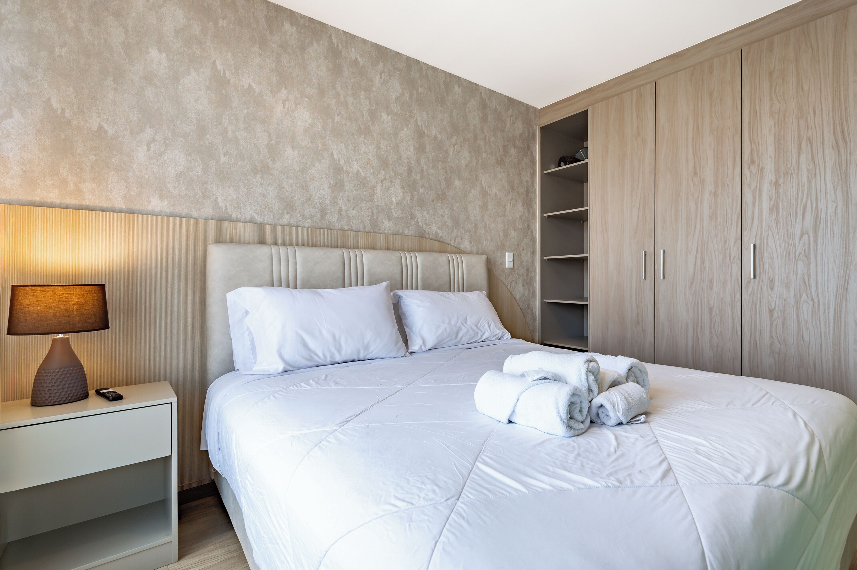 A neatly made bed with white linens, a bedside lamp, and a rolled towel on the bed.