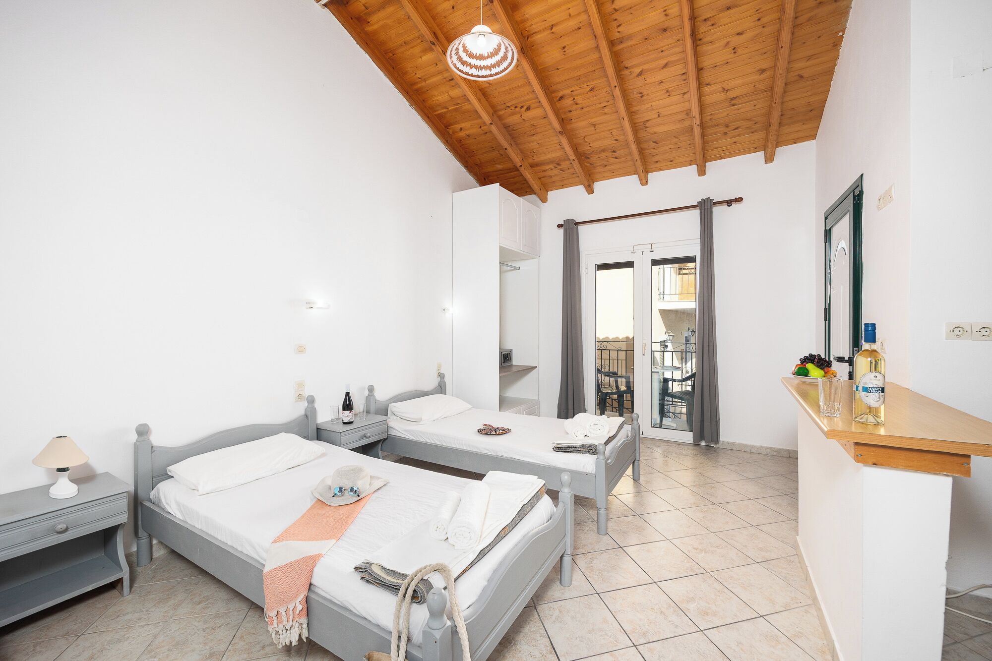 Une chambre avec deux lits, un plafond en bois, un petit bureau avec un miroir et une porte-fenêtre donnant sur un balcon.