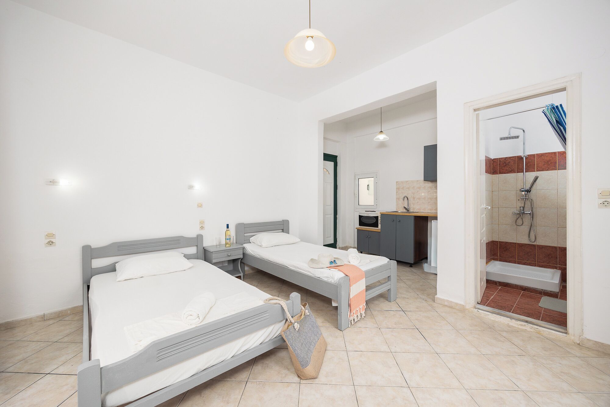 A modern hotel room with two single beds, a small desk, a mirror, and a shower area.
