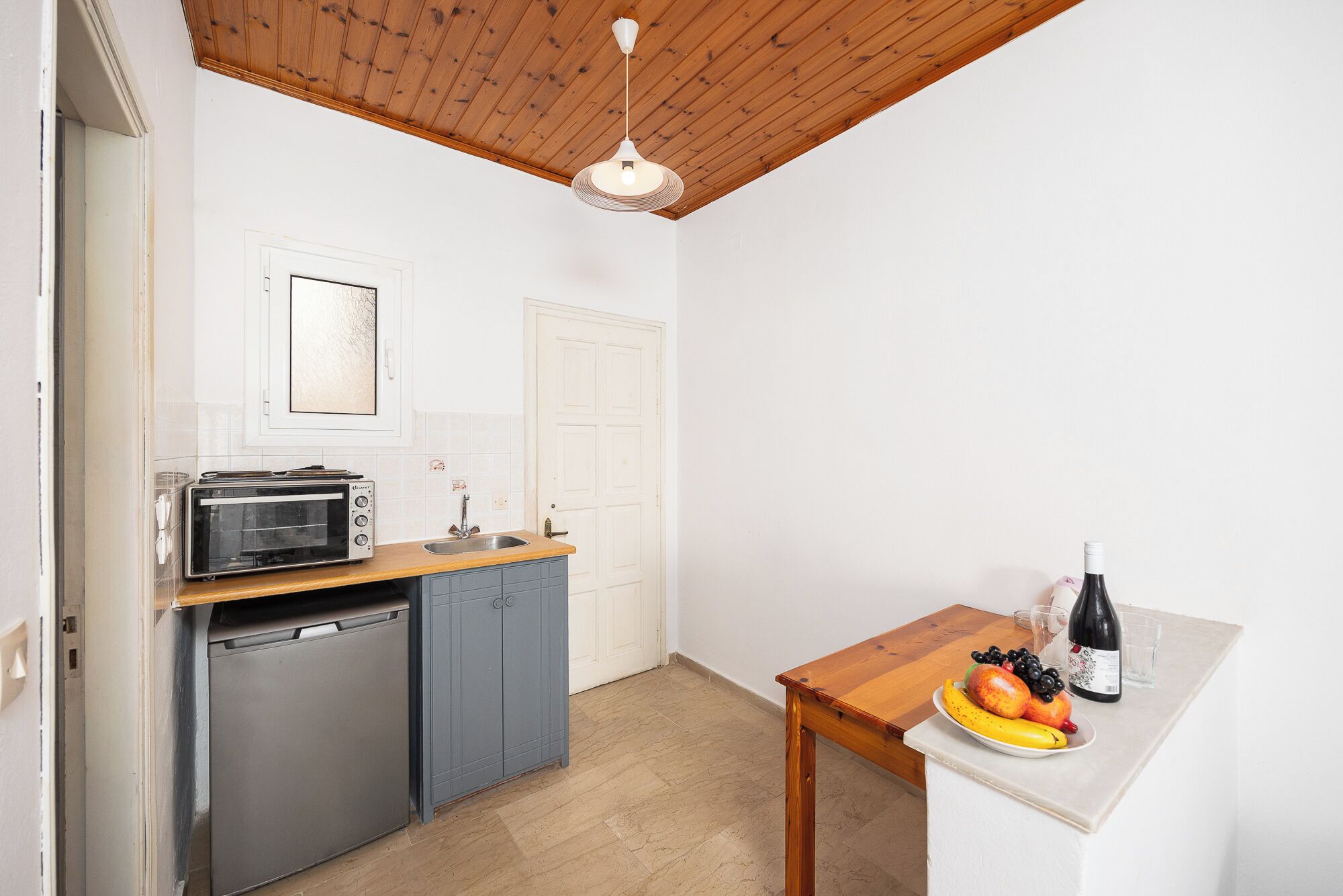 A compact kitchen with a built-in oven, a sink, and a small table with a bottle of wine and fruit.