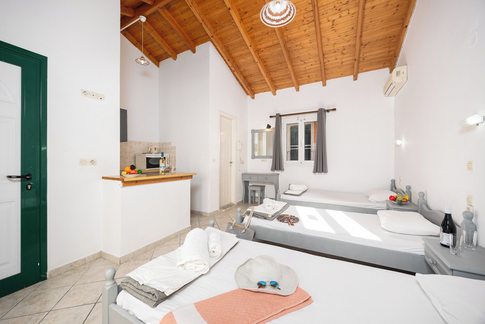 A small, clean room with a wooden ceiling, a bed, a bedside table, a mirror, and a small kitchenette.