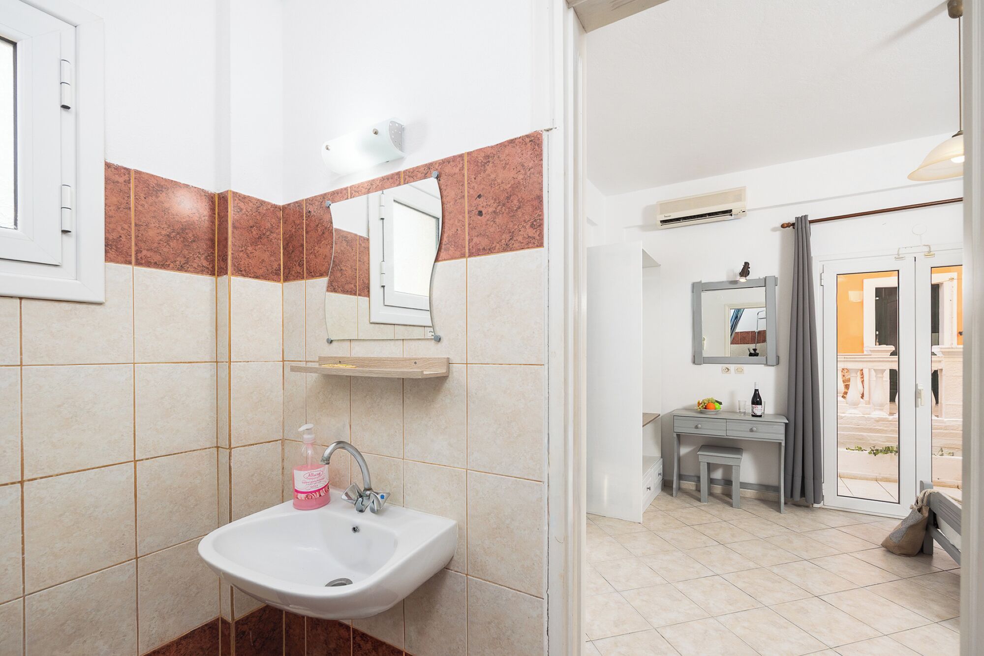 Une salle de bain avec un lavabo, un miroir et un porte-serviettes. On y trouve une commode avec un miroir et une bouteille de shampoing. La salle de bain est carrelée et possède une fenêtre.
