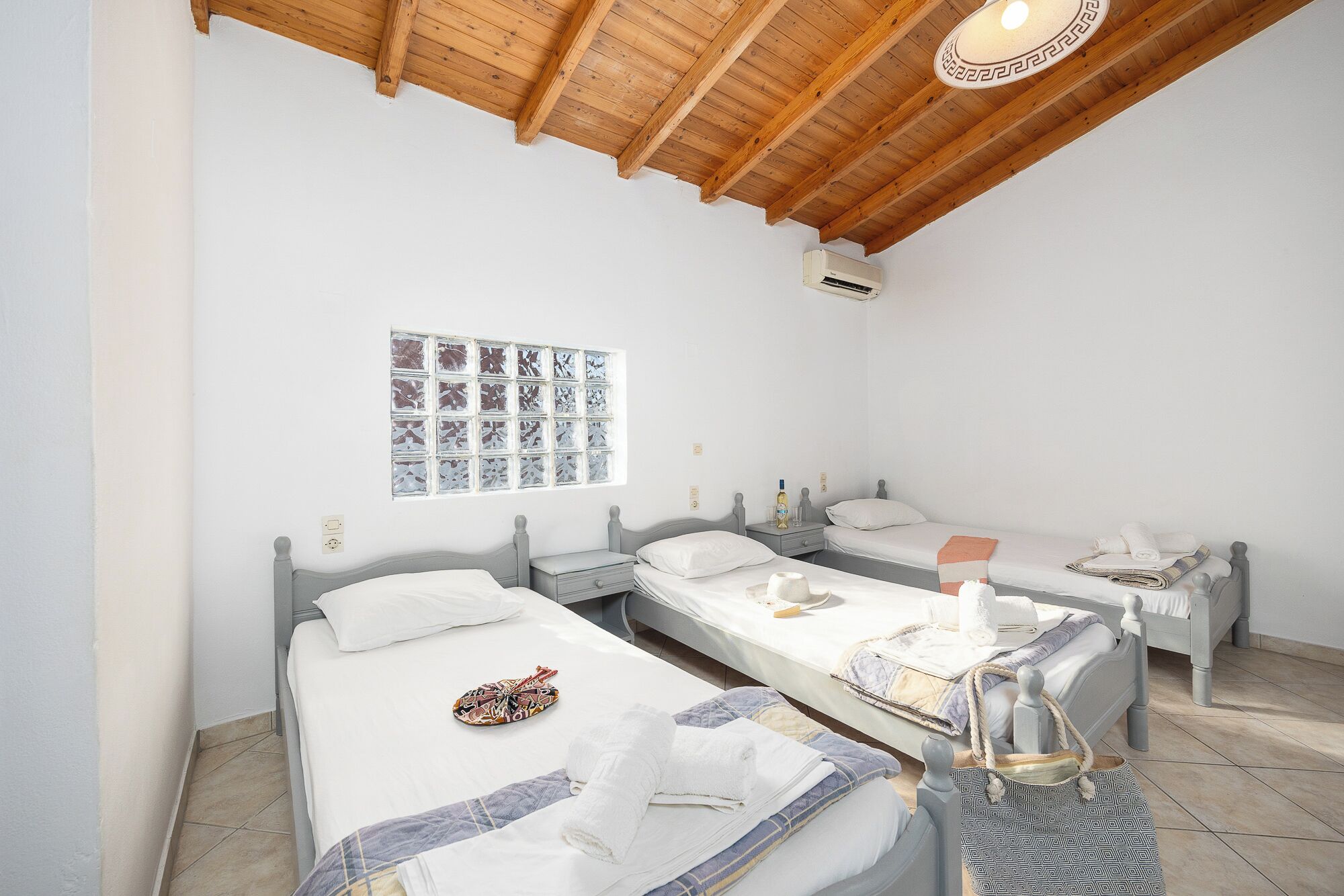 A room with three single beds, a wooden ceiling, an air conditioning unit, and a wall with a decorative window.