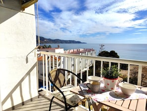 A balcony with a wicker chair, a table with a floral tablecloth, and a view of the sea.