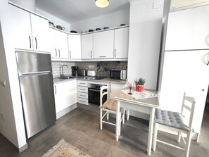 Cocina compacta con armarios blancos, refrigerador de acero inoxidable, microondas, tostadora y una mesa de comedor con sillas.