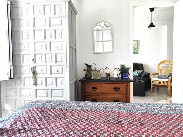 Una habitación con una colcha estampada, un aparador con objetos decorativos, una silla de mimbre y una puerta blanca con un ventanal.