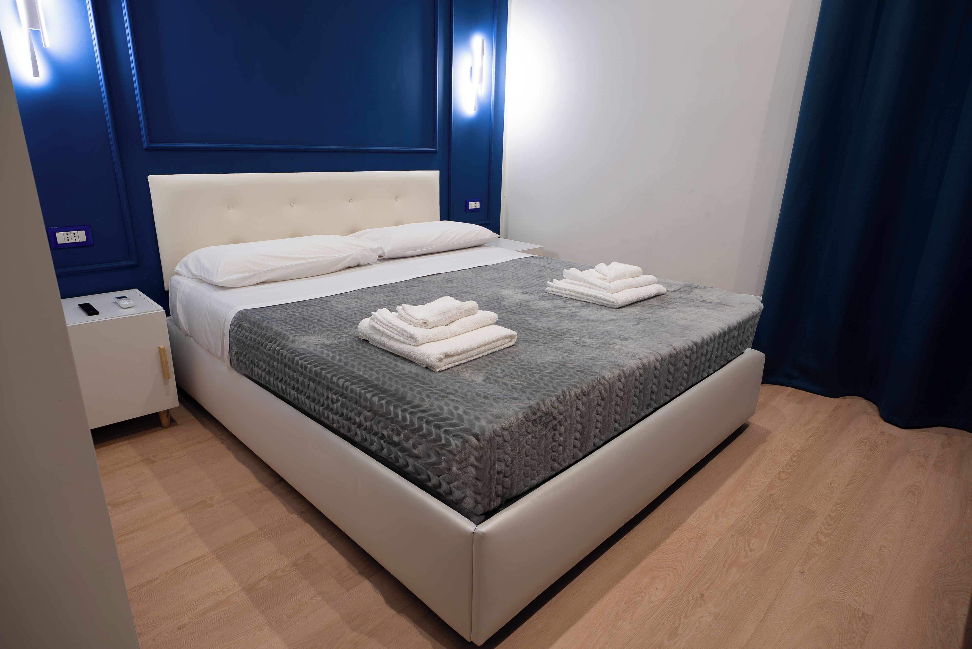 A neatly made bed with white linens and folded towels, a white nightstand with a remote control, and a blue wall with a light fixture.
