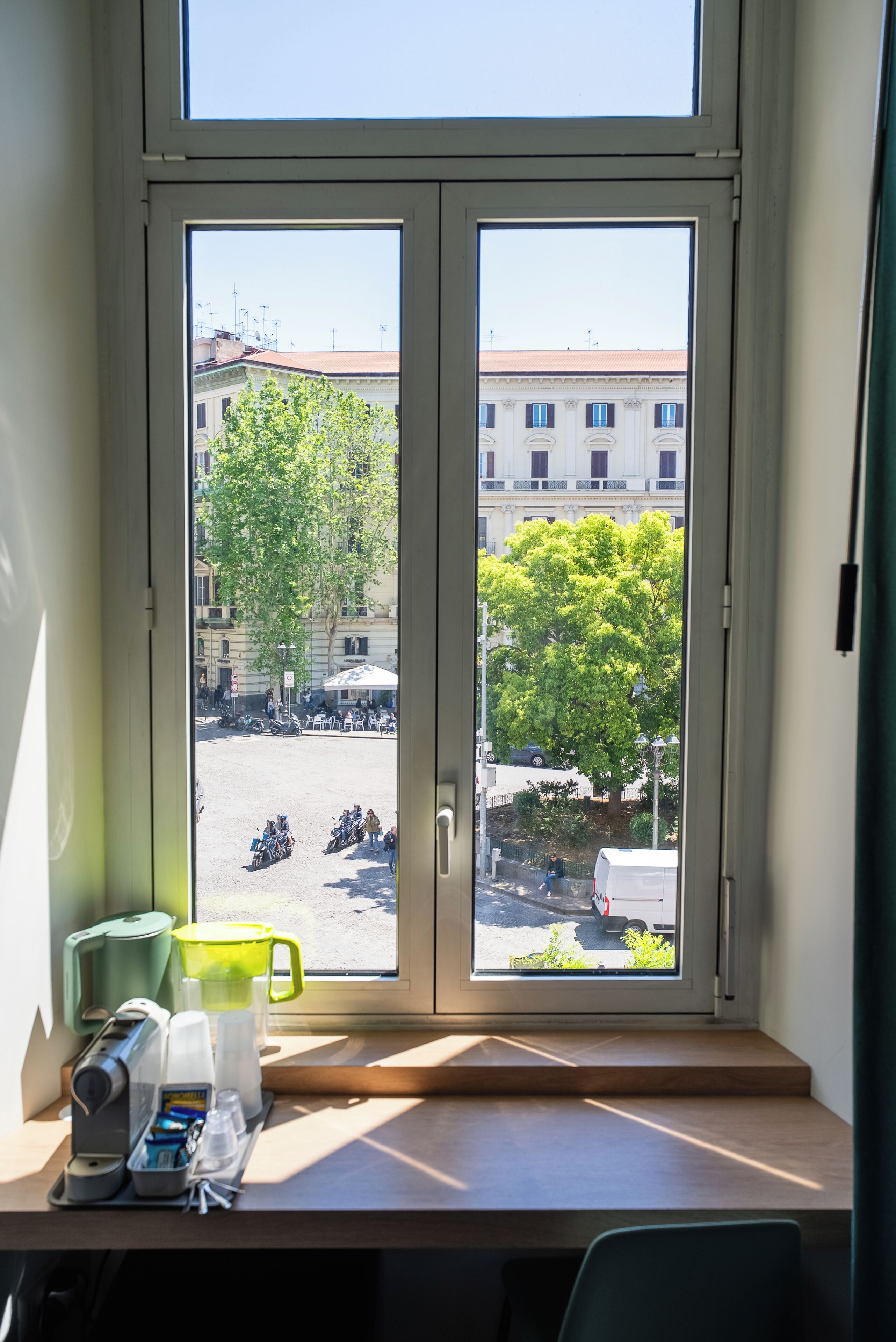 A window with a view of a street, a table with a coffee maker, and a green container.