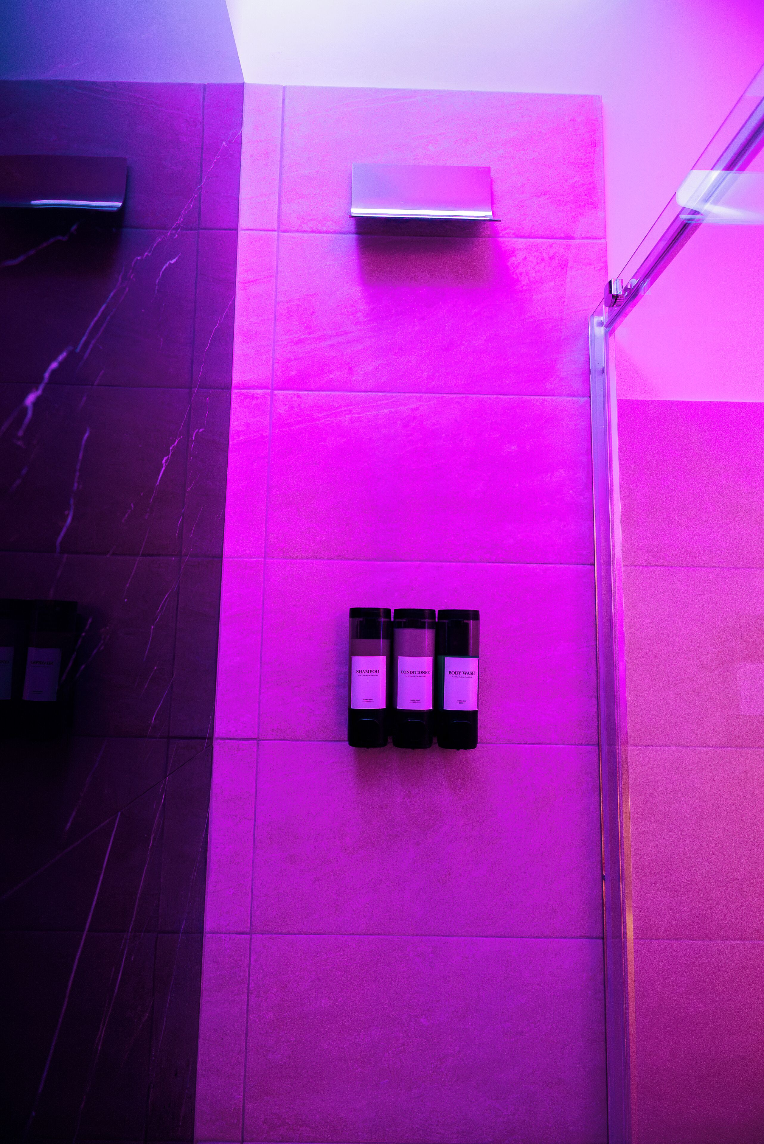 A bathroom with a purple-lit wall, a mirror, and three bottles of shampoo.