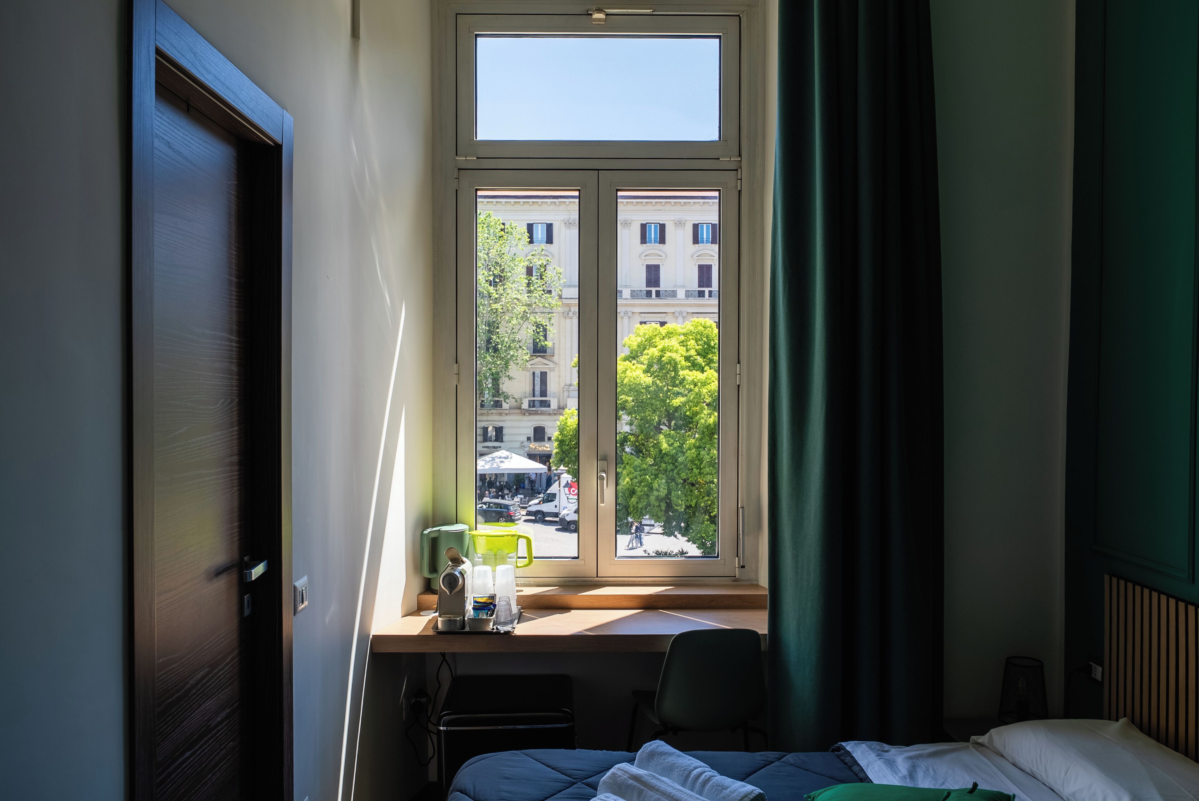 A hotel room with a bed, a desk with a chair, a window with a view of buildings and trees, and a door.