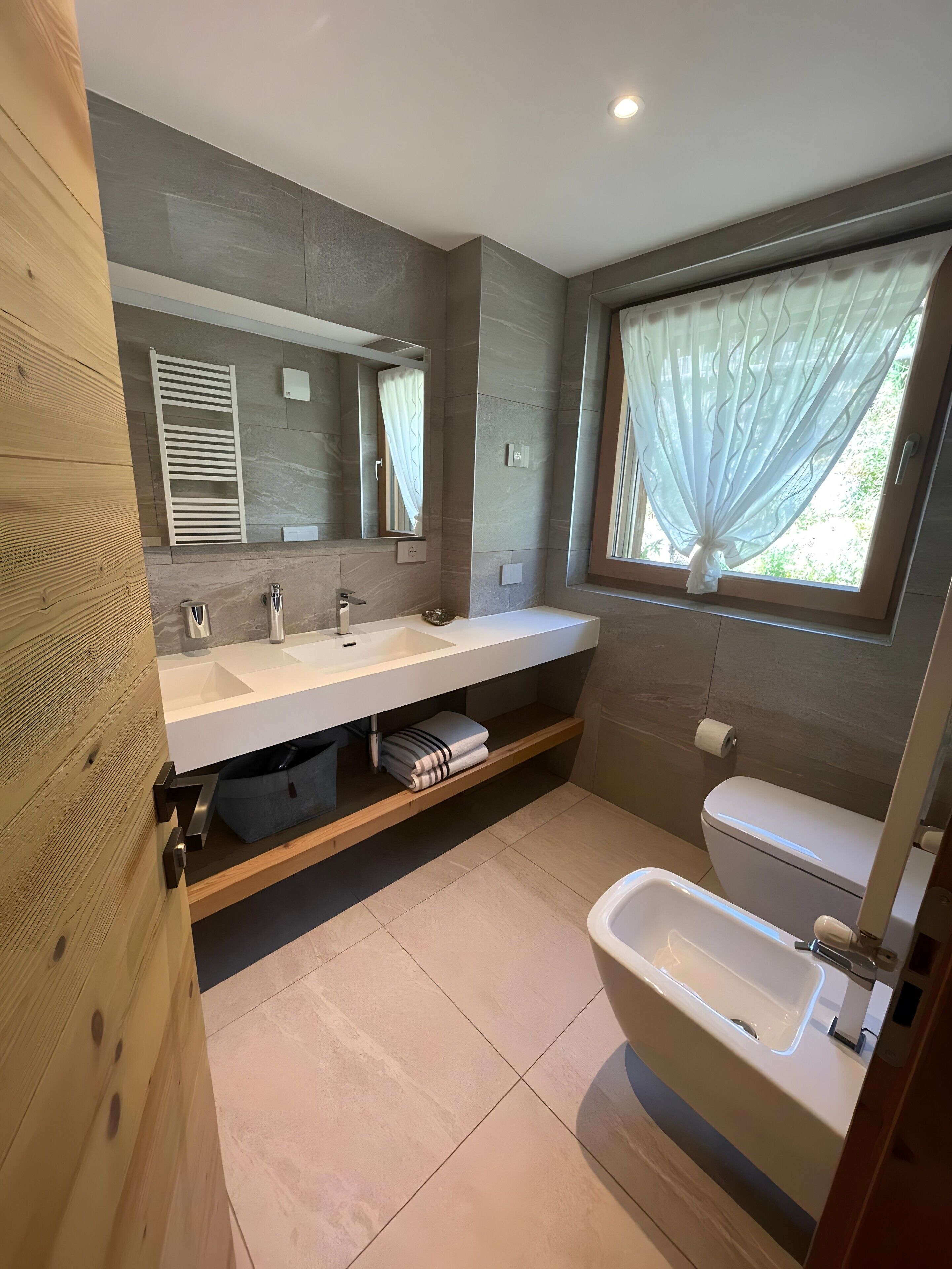 A modern bathroom with a wooden vanity, a large mirror, a dual sink, a toilet, and a bidet.