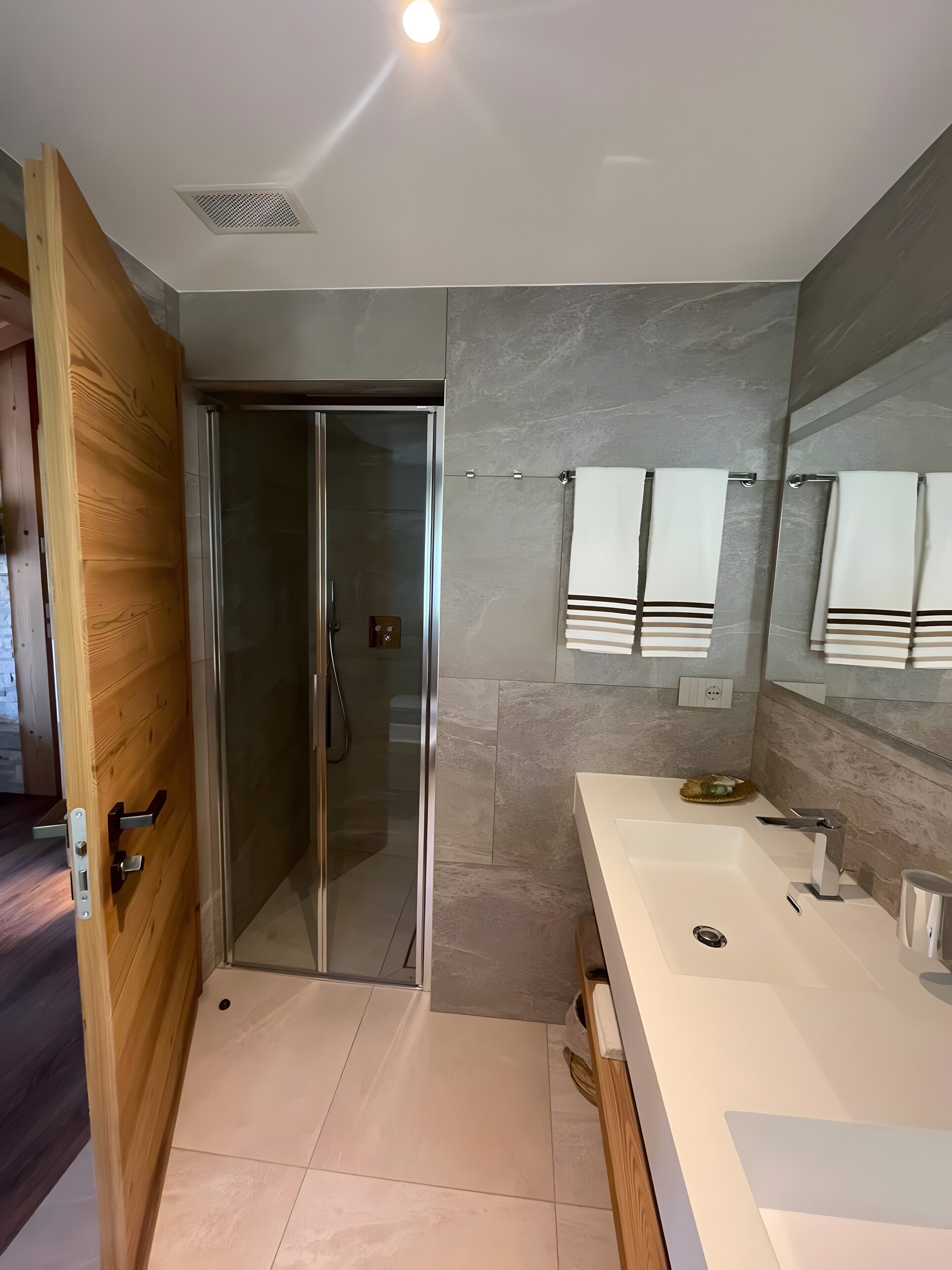 Une salle de bain moderne avec une douche à parois de verre, une porte en bois, un grand miroir et un lavabo blanc.