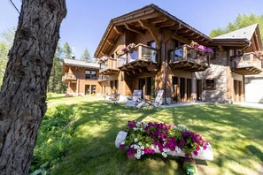 A wooden cabin with multiple balconies, a stone facade, and a well-maintained lawn with flower beds.