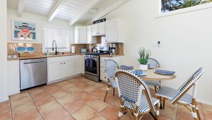 A kitchen with white cabinets, a stainless steel dishwasher, an oven, and a coffee maker. There are wicker chairs around a small table with a plant.