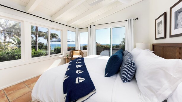 Una camera da letto con un letto grande, una ventola a soffitto e vista sull'oceano attraverso ampie finestre.