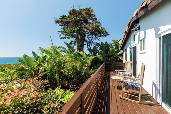 A wooden deck with a rocking chair and a table, surrounded by lush tropical plants and a large tree. - Charming Seaside Bungalows - 2BR + Studio - Ocean Views (Del Mar)