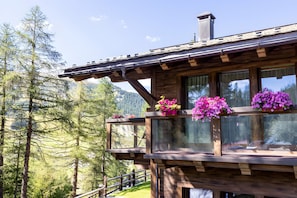 A wooden cabin with large windows, a balcony with flowers, and a view of a forested mountain area.