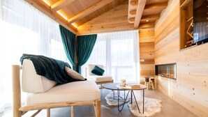 A cozy cabin interior with a sofa, a chair, a small round table, a fireplace, and a large window with curtains.