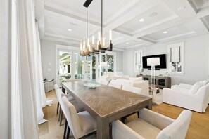 A modern dining area with a wooden table and chairs, a chandelier, and a view of the living room with white sofas and a TV.