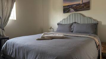Un lit bien fait, avec une couverture grise et des oreillers, une lampe de chevet et, au mur, un tableau représentant un phare.