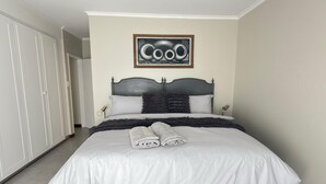 A neatly made bed with white linens and two rolled towels, a dark headboard, and a framed artwork above it.