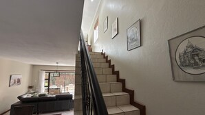 A staircase with a black railing leading to a landing. There are framed pictures on the walls, a sofa, and a dining table with chairs.